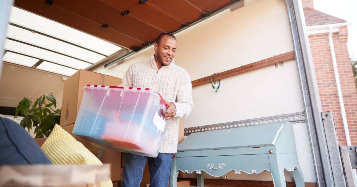 A man smiles and holds a clear moving box filled with items as he removes it from the back of an open moving truck.