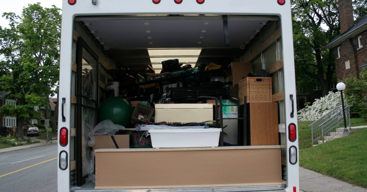 A white moving van with an open back gate and a ramp leading up to it parked on the side of the road near a building.