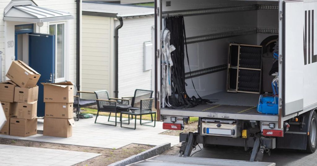 A large moving truck with an open back sits near a house with a blue door, surrounded by stacked cardboard boxes.