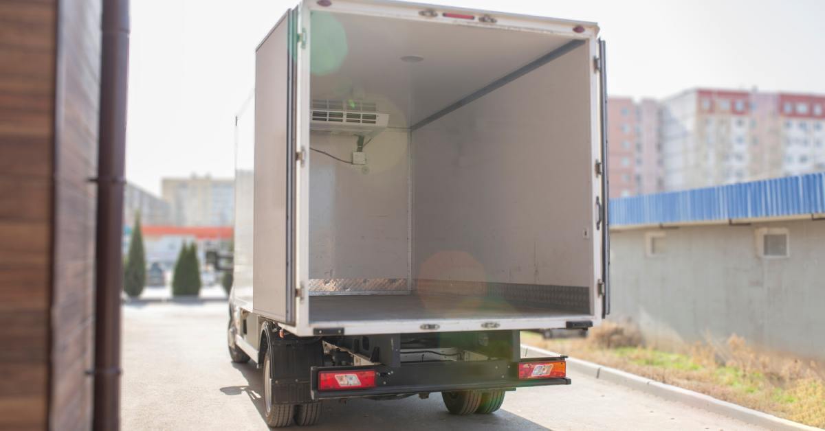 A back view of a parked white box truck with its rear doors open, showing an empty interior and ventilation system.