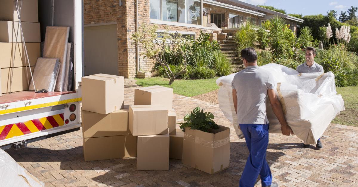 Two men carry a plastic-wrapped sofa toward a truck already loaded with boxes and household items on a sunny day.
