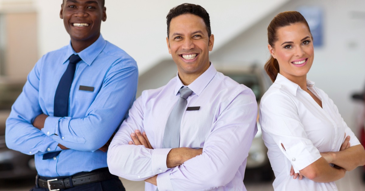 Three individuals in professional attire stand together while crossing their arms, with cars behind them.
