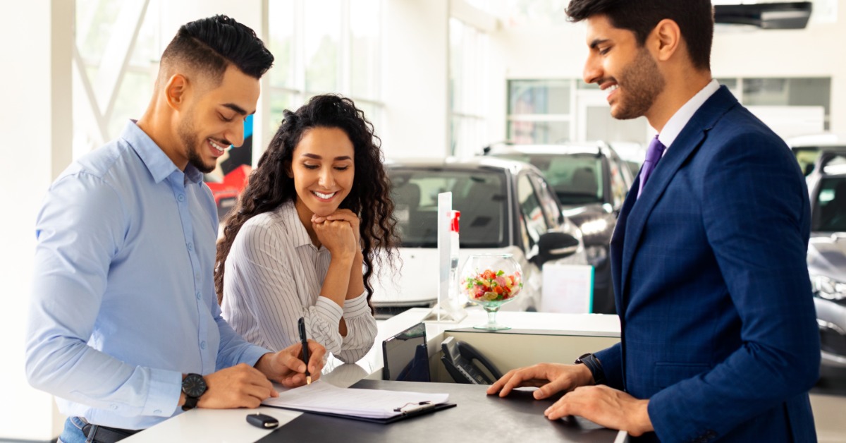 A man and a woman are signing papers at a checkout desk. Another man in a suit is on the opposite side of them, smiling.