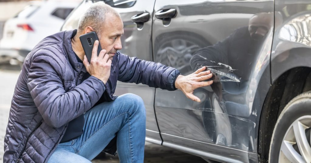A man standing beside a damaged car in a parking lot, talking on his phone after a collision with visible door damage.