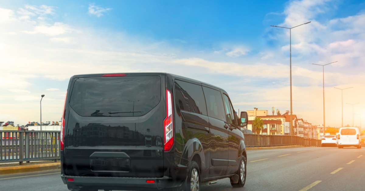 A black passenger van is driving on a highway during the day. There are several cars ahead of the van on the street.