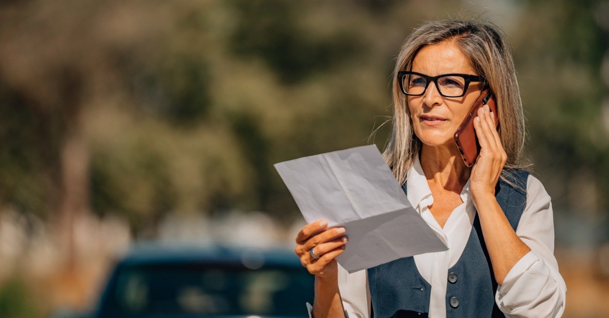 A woman wearing black glasses is holding documents while talking on a cellphone near a vehicle parked by trees.