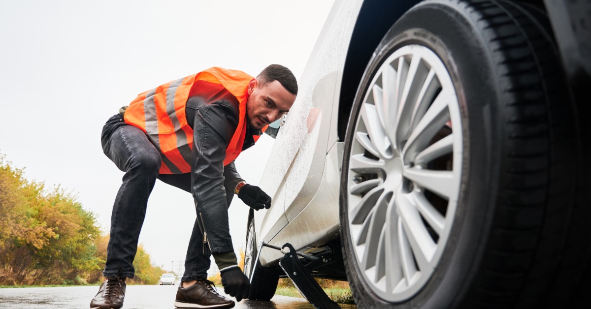 A person uses a car jack to lift a vehicle beside a wet road. They wear an orange safety vest and black gloves.