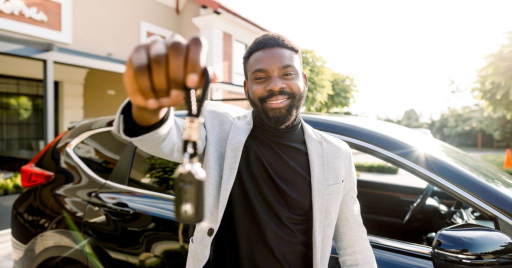 A Black man stands in front of an SUV with its front window rolled down. He smiles as he holds the car keys.