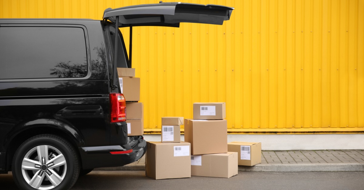 A black delivery van is parked beside a stack of boxes near a yellow building in an outdoor loading area.