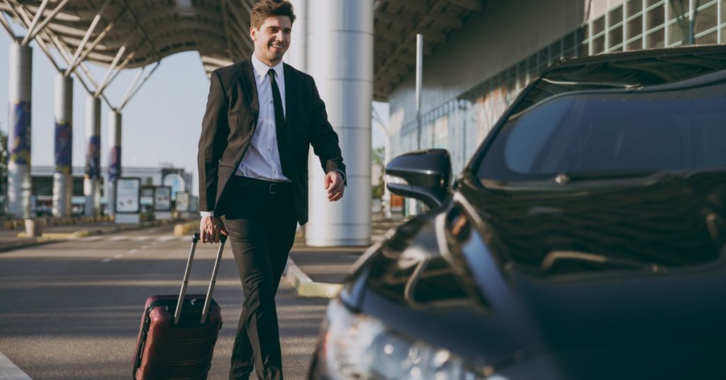 A tall Caucasian man in a suit pulling a suitcase toward a parked black car outside a covered airport terminal.