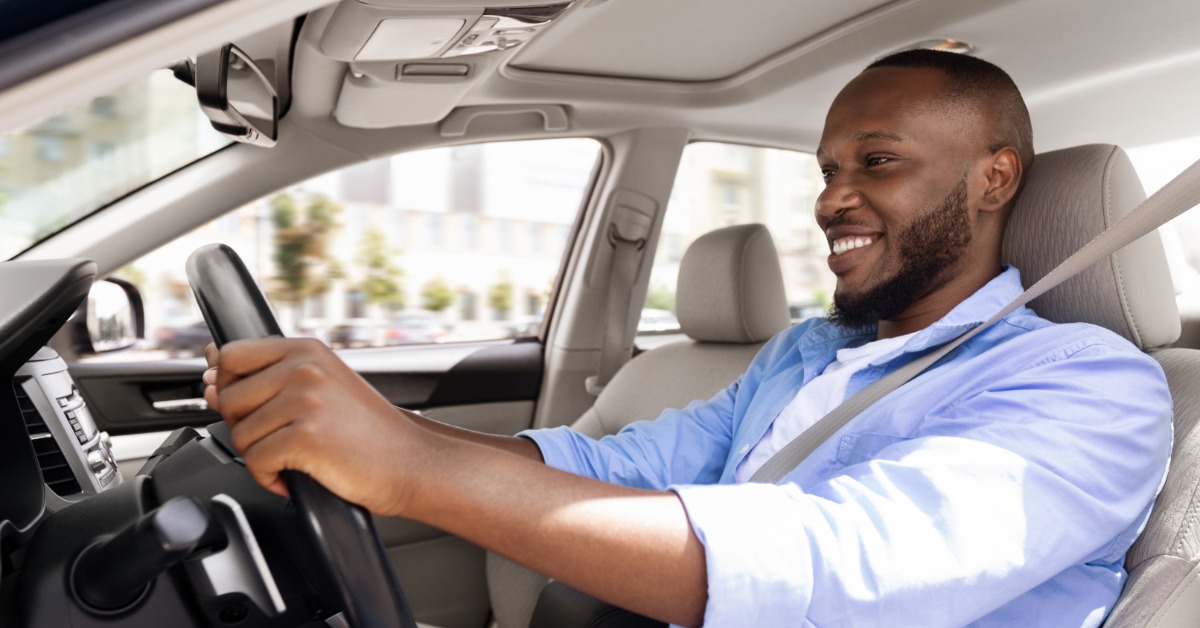 A Black man wearing a seat belt and a blue shirt smiles as he drives a car with a light interior through a city.