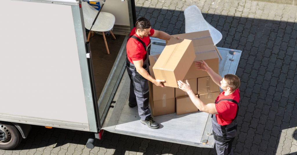 Two movers carry cardboard boxes from the back of a white truck, with more boxes stacked inside the cargo area.