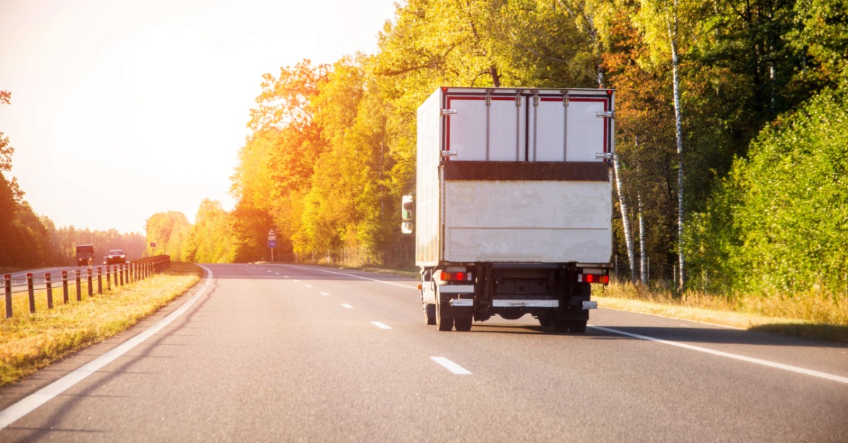A white refrigerated truck drives along a highway at sunset, with trees and a warm sky in the background.