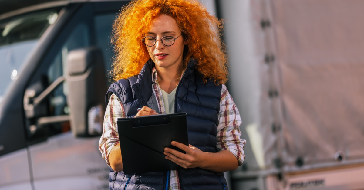 A woman with red hair writes notes on a black clipboard while standing in front of a large white truck outdoors.
